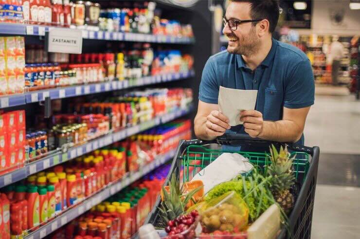 Uomo felice tra gli scaffali al supermercato con carrello e lista della spesa